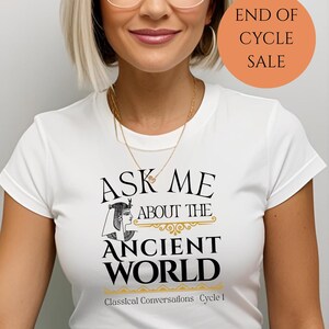 May include: White t-shirt with black text that reads "Ask Me About The Ancient World" with a stylized image of an Egyptian woman. Below the text is "Classical Conversations Cycle 1". The image has a peach colored circle with the text "End of Cycle Sale" in black.