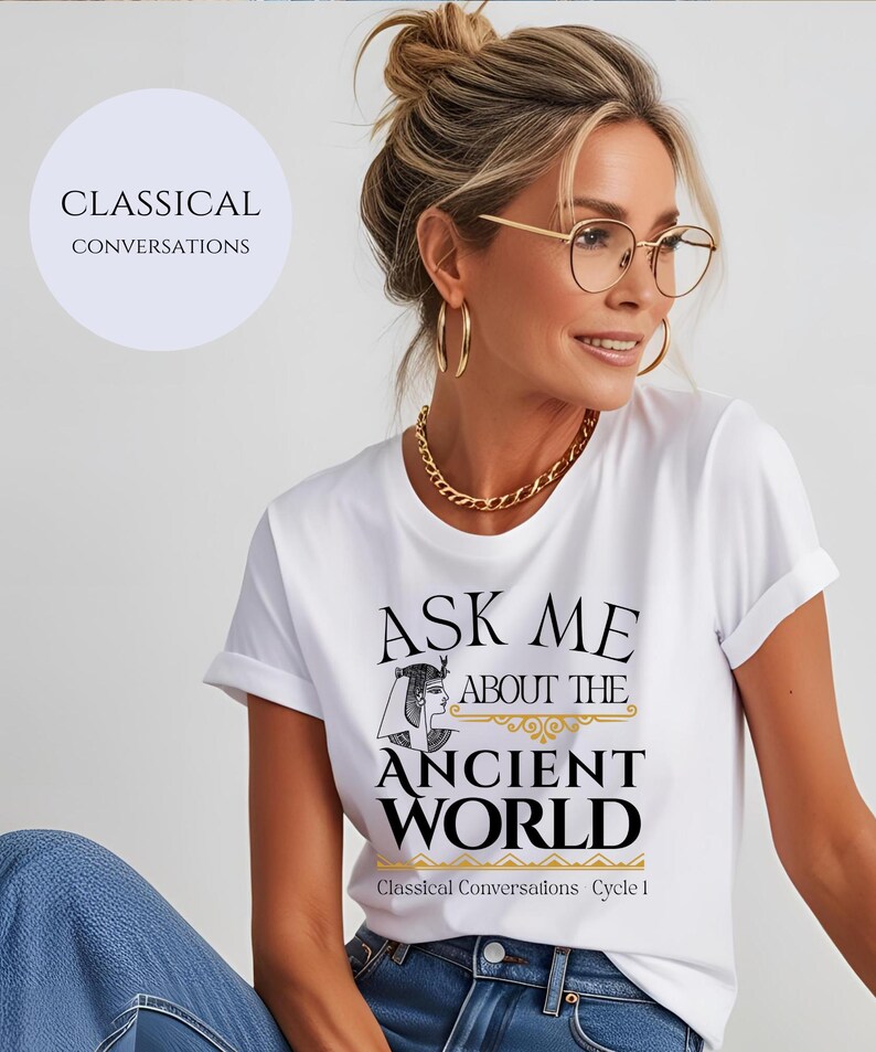 May include: White t-shirt with black text that reads "Ask me about the ancient world."  The text is surrounded by a gold design.  The text "Classical Conversations Cycle 1" is printed in gold at the bottom of the shirt.