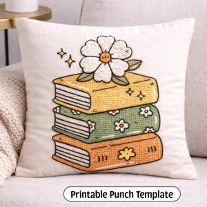 May include: A square off-white throw pillow featuring an embroidered design of a stack of three books topped with a large white flower. The top book is yellow, the middle is green with white flowers, and the bottom is orange with a yellow flower.