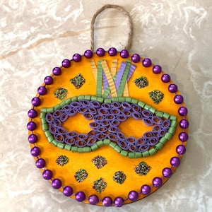 May include: A round, wooden Mardi Gras mask decoration. The mask features a purple eye mask design, green and purple accents, and is surrounded by purple beads. A jute loop is attached for hanging.