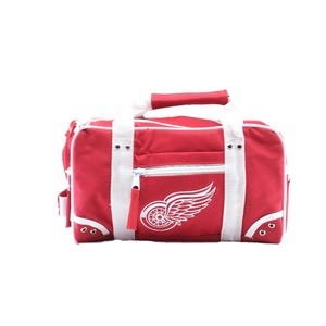 May include: Red duffel bag with white detailing and a zip. It has a white logo of a winged wheel on the front. Includes white handles and a shoulder strap. The bag is designed for carrying sports equipment or travel essentials.