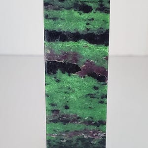 May include: A rectangular, green and black stone obelisk with a pointed top. The stone has a natural pattern of green, black, and purple hues. The obelisk is a decorative item.