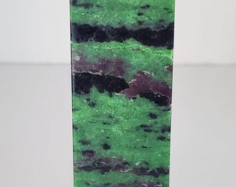Op de afbeelding: Een rechthoekige, groene en zwarte stenen obelisk met een puntige top. De steen heeft een natuurlijk patroon van groene, zwarte en paarse tinten. De obelisk is een decoratief item.