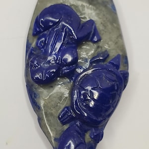 Puede incluir: Colgante ovalado tallado en piedra gris y azul, con un relieve detallado de una rana y una tortuga. La rana está colocada encima de la tortuga, ambas en un tono azul profundo. Cordón beige adjunto.