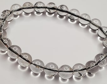 Könnte beinhalten: Armband mit klaren Quarz-Kristallperlen. Das Armband besteht aus runden, durchscheinenden Perlen mit dunklen Einschlüssen, die auf einer schwarzen elastischen Schnur aufgefädelt sind. Die Perlen haben einen Durchmesser von etwa 1 cm. Das Armband wird auf einer weißen Oberfläche präsentiert.