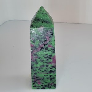 Puede incluir: Un cristal de zoisita rectangular pulido con una parte superior puntiaguda. La piedra presenta un patrón moteado de tonos verdes, negros y rosas. El cristal está de pie sobre un fondo blanco.