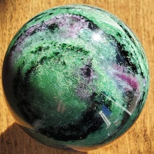 May include: A polished sphere of ruby in zoisite. The orb features a mix of green, black, and pink hues, with a glossy, reflective surface. The stone's colors are swirled and mottled, creating a unique pattern. The sphere is a decorative item.