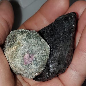 Puede incluir: Dos piedras preciosas en bruto se sostienen en una mano. Una es una roca rugosa de color verde oscuro con un centro rosa. La otra es una piedra grande, negra, de forma irregular. Las piedras preciosas tienen un aspecto natural y sin pulir.