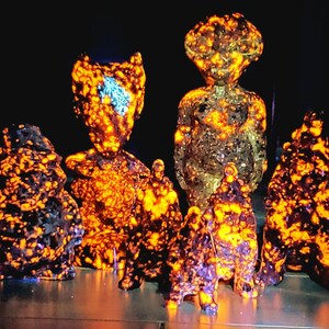 May include: A group of abstract sculptures with a speckled texture. The sculptures are predominantly dark with bright orange and yellow highlights, giving a glowing effect. The forms vary, including humanoid and amorphous shapes.