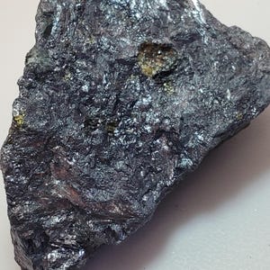 May include: A close-up of a dark gray mineral specimen with a rough, textured surface. The rock has a metallic sheen and small pockets of yellow and gold coloration. The overall shape is irregular and jagged.