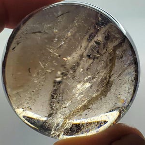 May include: A clear, round crystal ball with internal fractures and inclusions. The sphere has a smoky, light brown color with areas of transparency. The crystal is held in a hand, with the background out of focus.