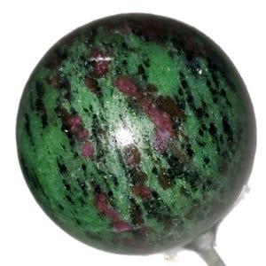 Könnte beinhalten: Eine polierte Kugel aus grünem Zoisit mit Rubin-Einschlüssen. Der Edelstein weist ein gesprenkeltes Muster aus tiefem Grün auf, das mit leuchtend roten und schwarzen Flecken durchsetzt ist. Die Kugel ist auf einem klaren, dünnen Stiel montiert.