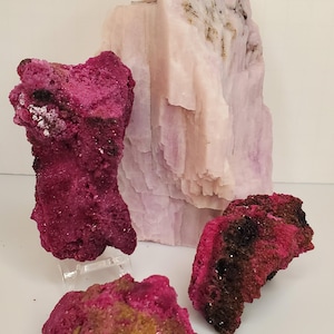 May include: A selection of raw mineral specimens. The image shows a large, pale pink, layered rock formation and several smaller, vivid magenta and dark red mineral pieces. The minerals have a rough, textured appearance.