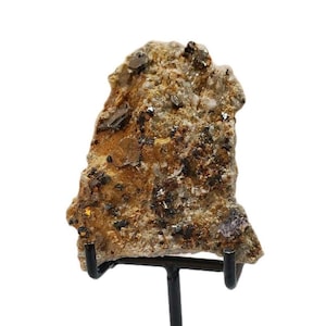 May include: A rough-hewn, natural mineral specimen with a mix of brown, tan, and gray tones. The rock features visible crystalline structures and metallic flecks, mounted on a black metal display stand. The overall texture is coarse and irregular.