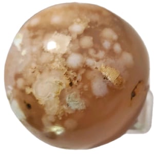 May include: A polished, round gemstone with a warm, earthy color palette. The stone features a blend of tan, cream, and gold hues, with a marbled appearance. The surface has a smooth, glossy finish, and the gemstone is set on a clear base.