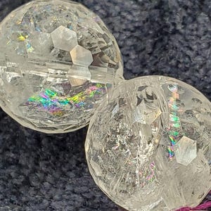May include: Two clear, faceted crystal spheres with iridescent rainbow reflections. The spheres have a cracked, geometric pattern, and are set against a dark blue background. The spheres are likely decorative or jewelry components.