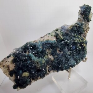 May include: A natural mineral specimen featuring a mix of colors. The piece displays a base of clear quartz with areas of deep teal and green crystal formations. The mineral is mounted on a clear stand.