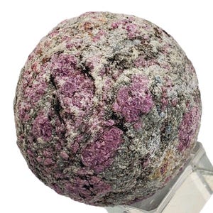 Könnte beinhalten: Ein kugelförmiger Edelstein mit rauer, strukturierter Oberfläche. Der Stein ist hauptsächlich tiefrosa gefärbt, mit grauen und weißen Flecken. Die Gesamtform ist rund, und es scheint sich um ein natürliches Mineralexemplar zu handeln.