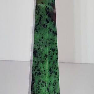 May include: A tall, polished stone obelisk with a pointed top. The stone features a vibrant green base color with black speckles and patches of deep red. The obelisk has a geometric, tapered shape.