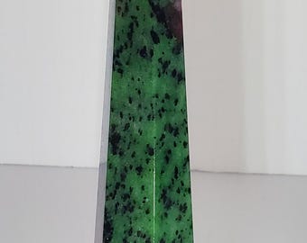 Op de afbeelding: Een gepolijste stenen obelisk met een puntige top. De steen heeft een levendige groene basiskleur met zwarte spikkels en vlekken van dieprood. De obelisk heeft een geometrische, taps toelopende vorm.