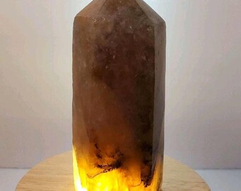 May include: A tall, brown crystal point with a faceted top. The base of the crystal glows with a warm, yellow light, creating a striking contrast with the darker upper portion. The crystal sits on a round, wooden base.