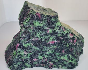 Op de afbeelding: Een grote, ruwe rots met een gevlekt uiterlijk. De steen heeft een mix van kleuren, waaronder diep groen, zwart en vlekken roze en rood. De rots heeft een onregelmatige vorm en een getextureerd oppervlak.