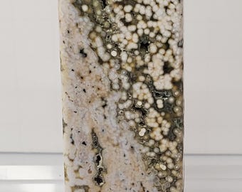 May include: A tall, polished gemstone obelisk. The stone features a speckled pattern of white, cream, and dark green, with a pointed top. The obelisk is a decorative item, likely for display or collection.