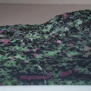 May include: A roughly cut stone with a mix of colours. The stone has a base of black and green with red and pink spots throughout. The stone is rectangular and has a slightly uneven top.