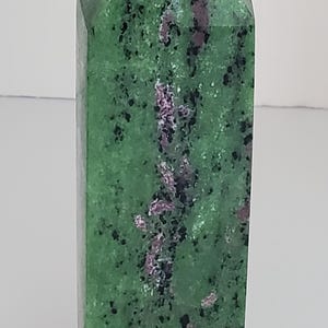 May include: A green and pink ruby zoisite crystal tower. The polished stone has a rectangular shape with a pointed top. The green base color is speckled with black and pink inclusions.