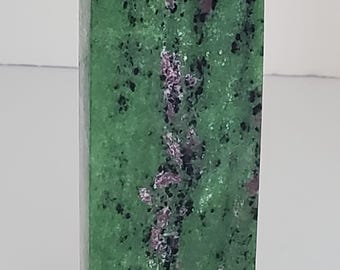 Op de afbeelding: Een groene en roze robijn-zoisiet kristallen toren. De gepolijste steen heeft een rechthoekige vorm met een puntige top. De groene basiskleur is bespikkeld met zwarte en roze insluitsels.