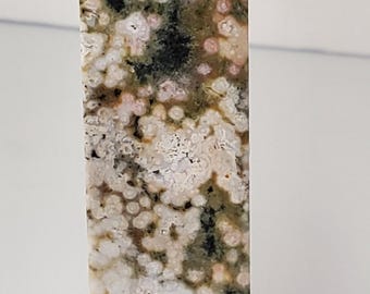 May include: A tall, rectangular crystal obelisk with a pointed top. The stone features a speckled pattern of white, pink, green, and black. The polished surface reflects light, highlighting the natural variations in the stone.