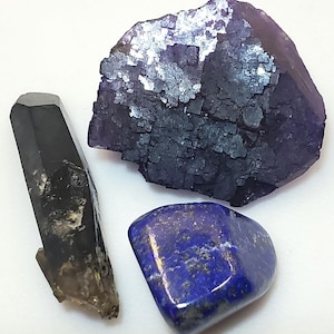 Może przedstawiać: Asortyment surowych kamieni szlachetnych, w tym ciemny kryształ kwarcu dymnego, duży fioletowy klaster fluorytu i polerowany niebieski kamień lapis lazuli. Kamienie szlachetne są prezentowane na białej powierzchni.
