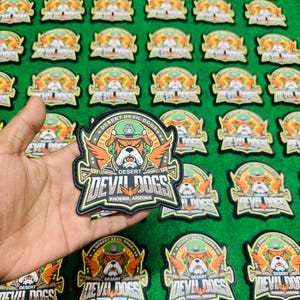May include: Embroidered patches with a military-inspired design. Each patch displays a bulldog in a helmet and wings, with the text "DESERT DEVIL DOGS" and "PHOENIX, ARIZONA". The patches are in black, green, orange, and white, arranged on a green surface.