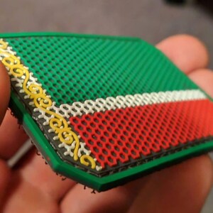 Pu&ograve; includere: Una toppa verde, rossa e bianca con un bordo decorativo giallo. La toppa ha uno sfondo a rete verde e un design a strisce rosse e bianche. La toppa &egrave; tenuta in una mano.