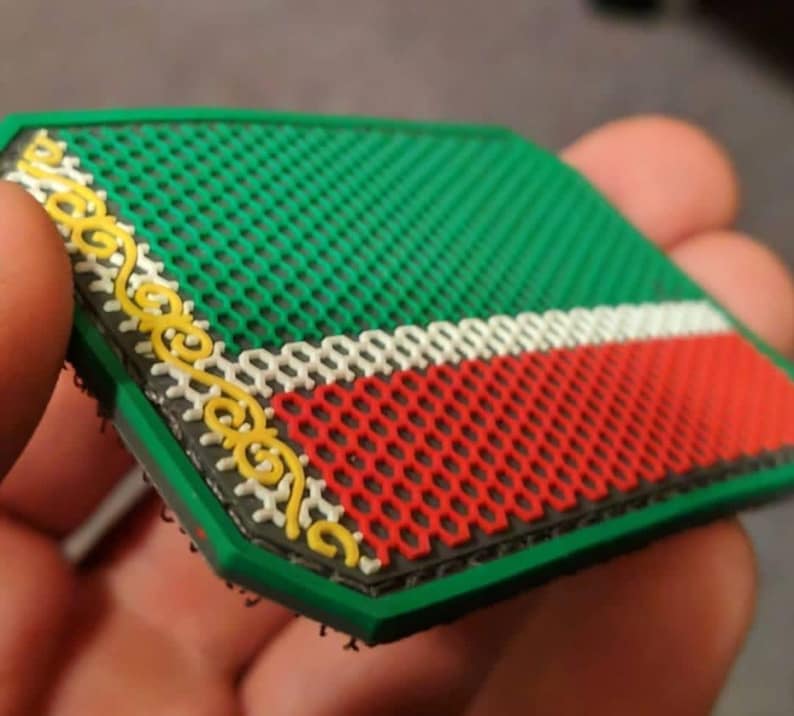 Pu&ograve; includere: Una toppa verde, rossa e bianca con un bordo decorativo giallo. La toppa ha uno sfondo a rete verde e un design a strisce rosse e bianche. La toppa &egrave; tenuta in una mano.