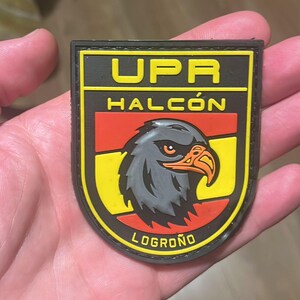 May include: A black, yellow, and red patch featuring the text "UPR HALC&Oacute;N LOGRO&Ntilde;O" and a detailed illustration of a hawk's head. The patch has a black border and is held in a hand.