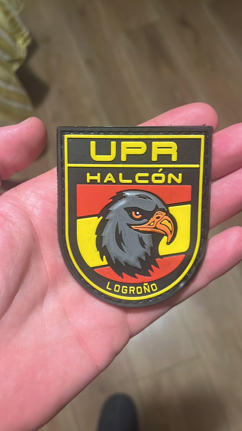 May include: A black, yellow, and red patch featuring the text "UPR HALC&Oacute;N LOGRO&Ntilde;O" and a detailed illustration of a hawk's head. The patch has a black border and is held in a hand.