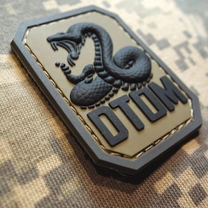 May include: A rectangular patch featuring a black snake design and the letters "DTOM" in black. The patch has a tan background with a black border, set against a camouflage pattern.