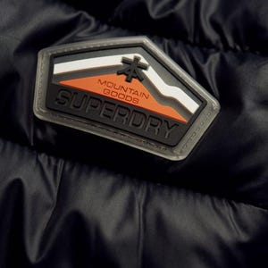 May include: Close-up of a black jacket featuring a Superdry patch. The patch is a geometric shape with a mountain design in white, orange, and black, and the words "MOUNTAIN GOODS" and "SUPERDRY".