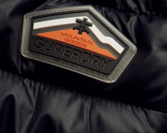 May include: Close-up of a black jacket with a Superdry patch. The patch is a geometric shape with a mountain design in white, orange, and black, and the words "MOUNTAIN GOODS" and "SUPERDRY".