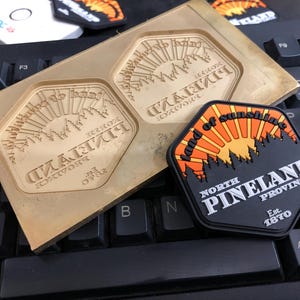 May include: A black and orange patch with the text "Land of Sunshine" and "North Pineland Province Est. 1870". A brass mold with the same design is also visible. The items are on a keyboard.