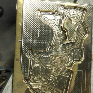 Pu&ograve; includere: Uno stampo metallico rettangolare color oro con un design dettagliato in rilievo. Il design sembra essere un'illustrazione complessa e stilizzata con elementi geometrici e meccanici. Lo stampo ha una superficie lucida e riflettente.