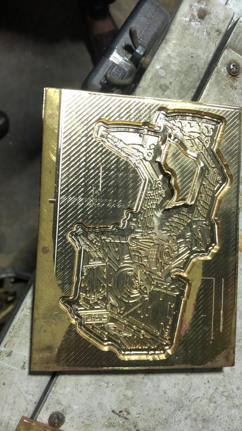 Pu&ograve; includere: Uno stampo metallico rettangolare color oro con un design dettagliato in rilievo. Il design sembra essere un'illustrazione complessa e stilizzata con elementi geometrici e meccanici. Lo stampo ha una superficie lucida e riflettente.