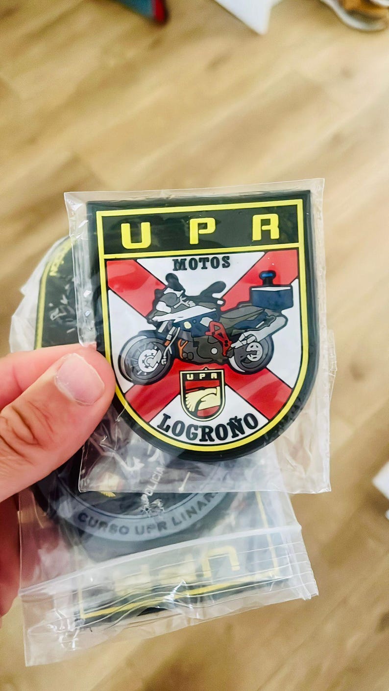 May include: A patch featuring a motorcycle graphic with the text "UPR MOTOS LOGRO&Ntilde;O". The patch has a black border, a red and white background, and the text "UPR" in yellow.