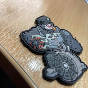 Pu&ograve; includere: Una toppa grigia e nera raffigurante un personaggio gorilla con un sigaro, che tiene in mano una grossa arma. La toppa ha un design dettagliato con accenti arancioni e grigi e un bordo nero. La toppa &egrave; su una superficie di legno.