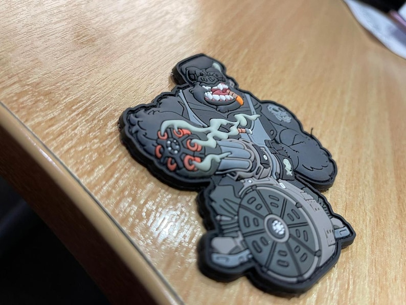 Pu&ograve; includere: Una toppa grigia e nera raffigurante un personaggio gorilla con un sigaro, che tiene in mano una grossa arma. La toppa ha un design dettagliato con accenti arancioni e grigi e un bordo nero. La toppa &egrave; su una superficie di legno.