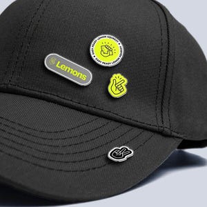 May include: Black baseball cap with multiple enamel pins attached. The pins display the word "Lemons", a hand gesture, and the phrase "Easy Peasy Lemon Squeezy". The pins are in yellow, black, and white.