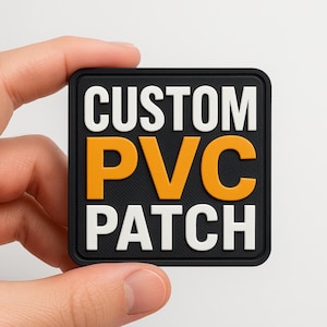 May include: A square black PVC patch with the words "CUSTOM PVC PATCH" in white and orange lettering. The patch is held in a hand against a white background. The patch has a textured surface.