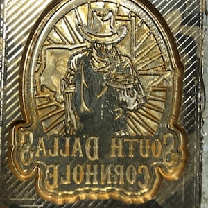 Pu&ograve; includere: Un timbro in metallo color oro con un'incisione dettagliata di una figura di cowboy con cappello, circondata da un disegno circolare. Il testo "SOUTH DALLAS" e "CORPORATION" &egrave; visibile.