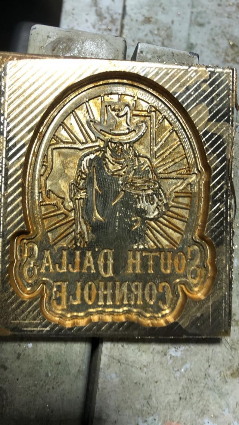 Pu&ograve; includere: Un timbro in metallo color oro con un'incisione dettagliata di una figura di cowboy con cappello, circondata da un disegno circolare. Il testo "SOUTH DALLAS" e "CORPORATION" &egrave; visibile.